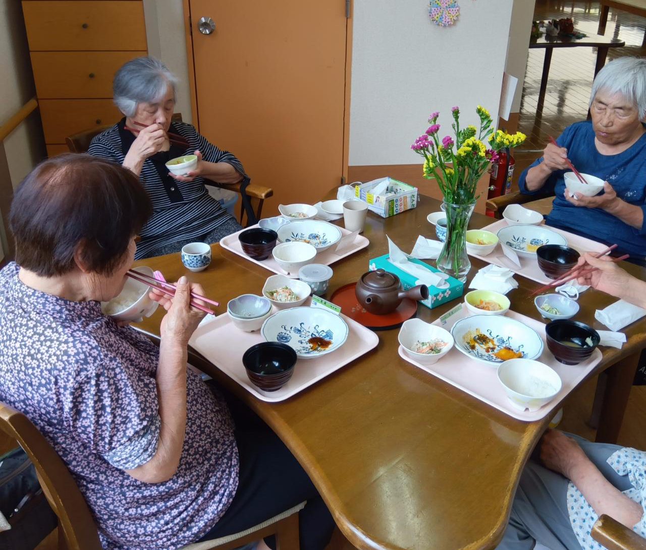 介護施設の食堂で複数の高齢入居者が食事をしている風景。花が飾られた温かみのある空間。